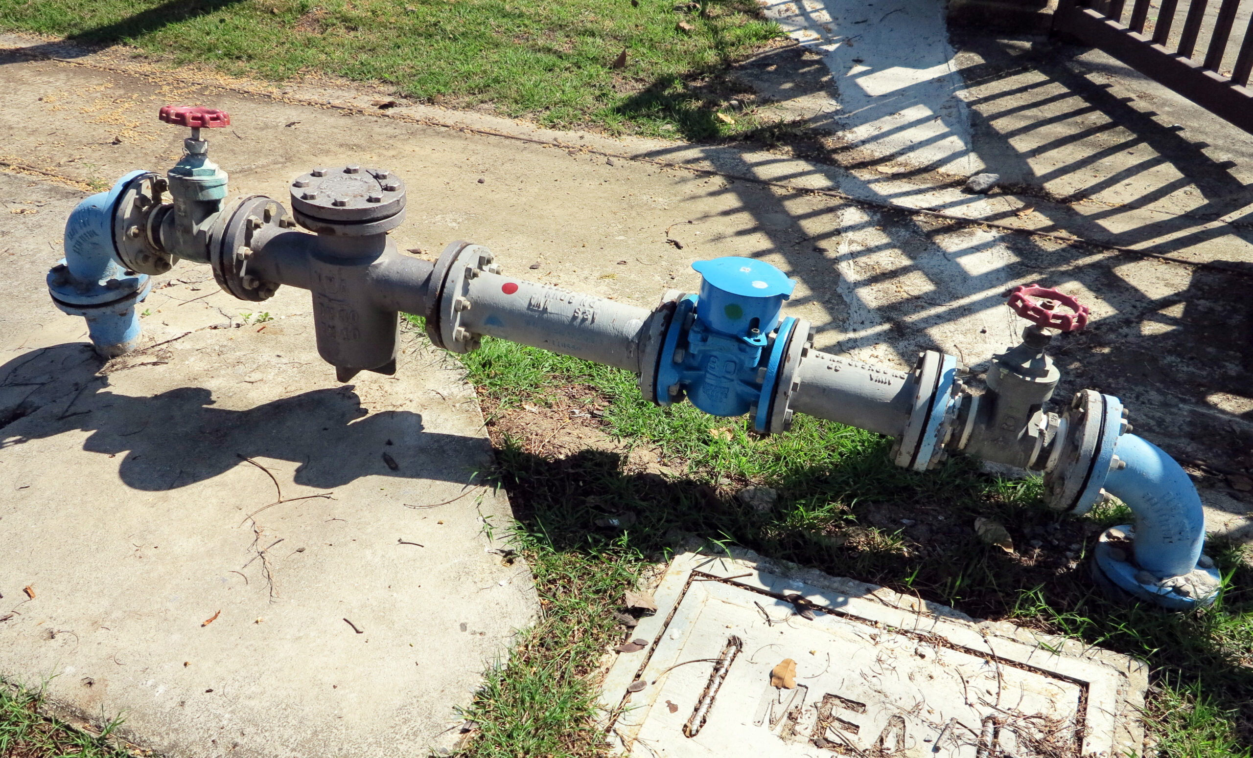 Water control pipeline with red and blue valves, mounted on a concrete base, surrounded by grass, possibly indicating water supply infrastructure.