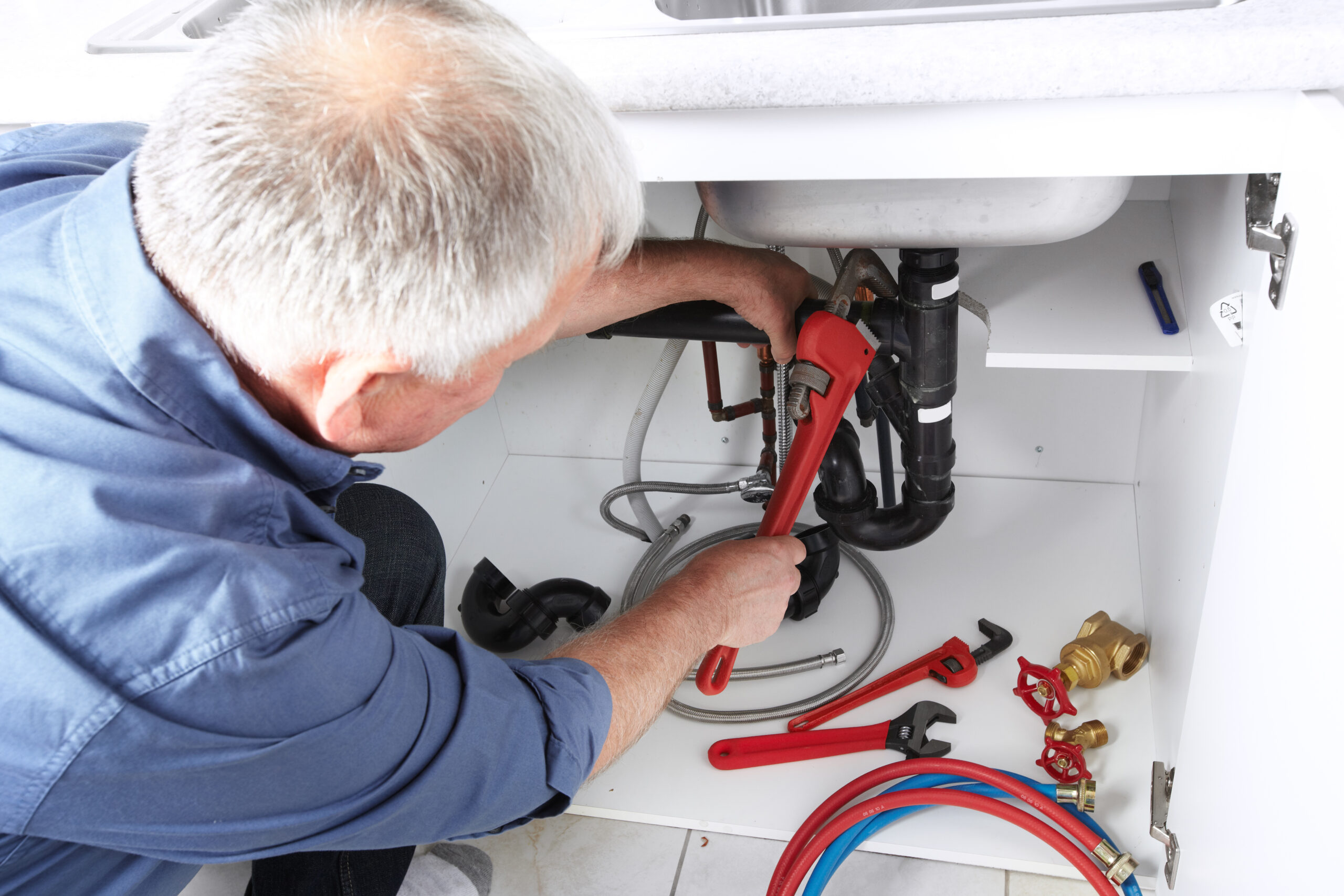 A plumber repairs pipes under a kitchen sink, using a red wrench. Various plumbing tools and fittings are visible, highlighting DIY maintenance.