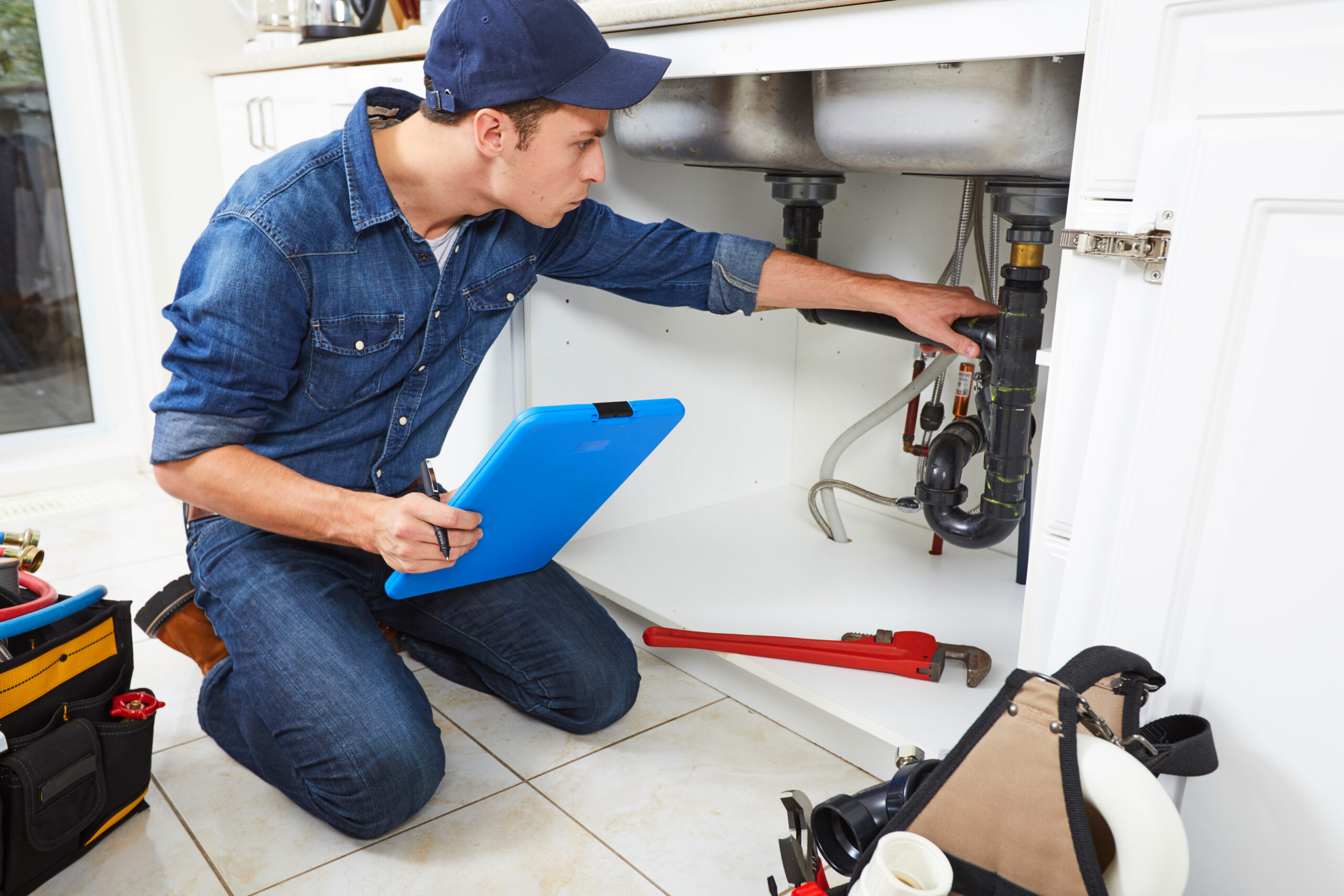 A plumber kneels in front of a kitchen sink, examining pipes while taking notes. Tools and equipment lie organized nearby, highlighting a service call.