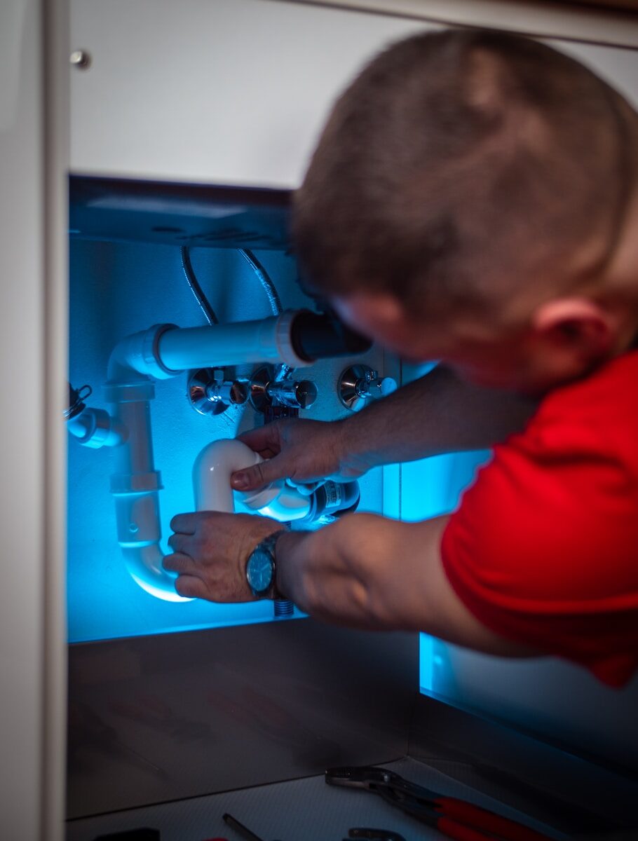 A plumber in a red shirt adjusts white pipes in a brightly lit under-sink area, showcasing plumbing repair work.